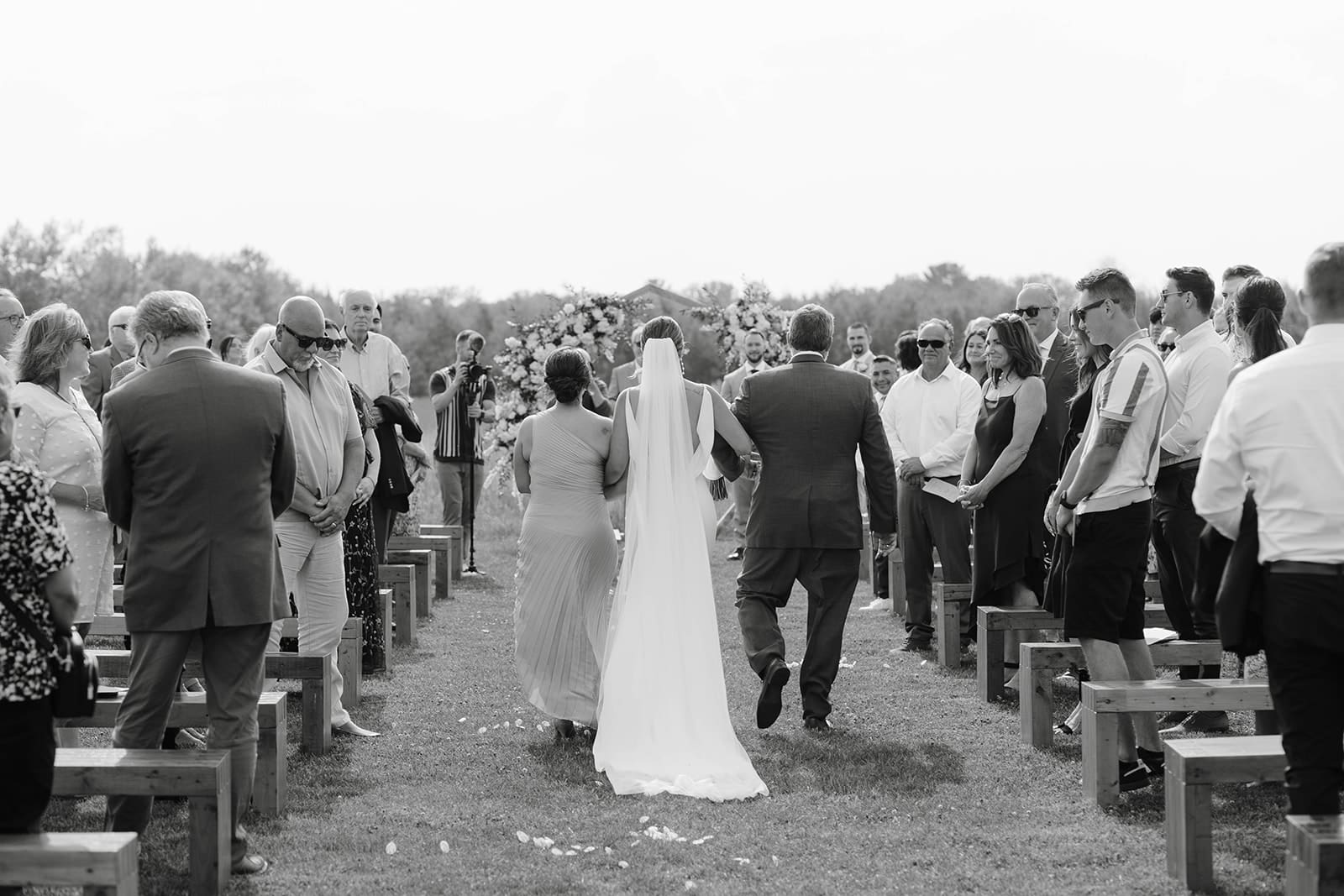 A photo of the back of a dress as the bride walks down the aisle at an Ivory North wedding in the summer. 