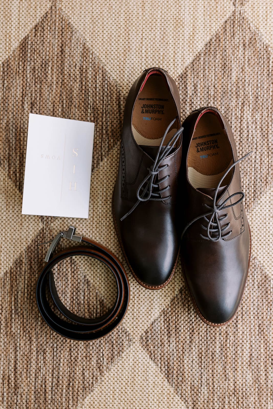 Flat lay details of the groom's details, including the shoes, belt and vows, on top of a diamond patterned rug. 