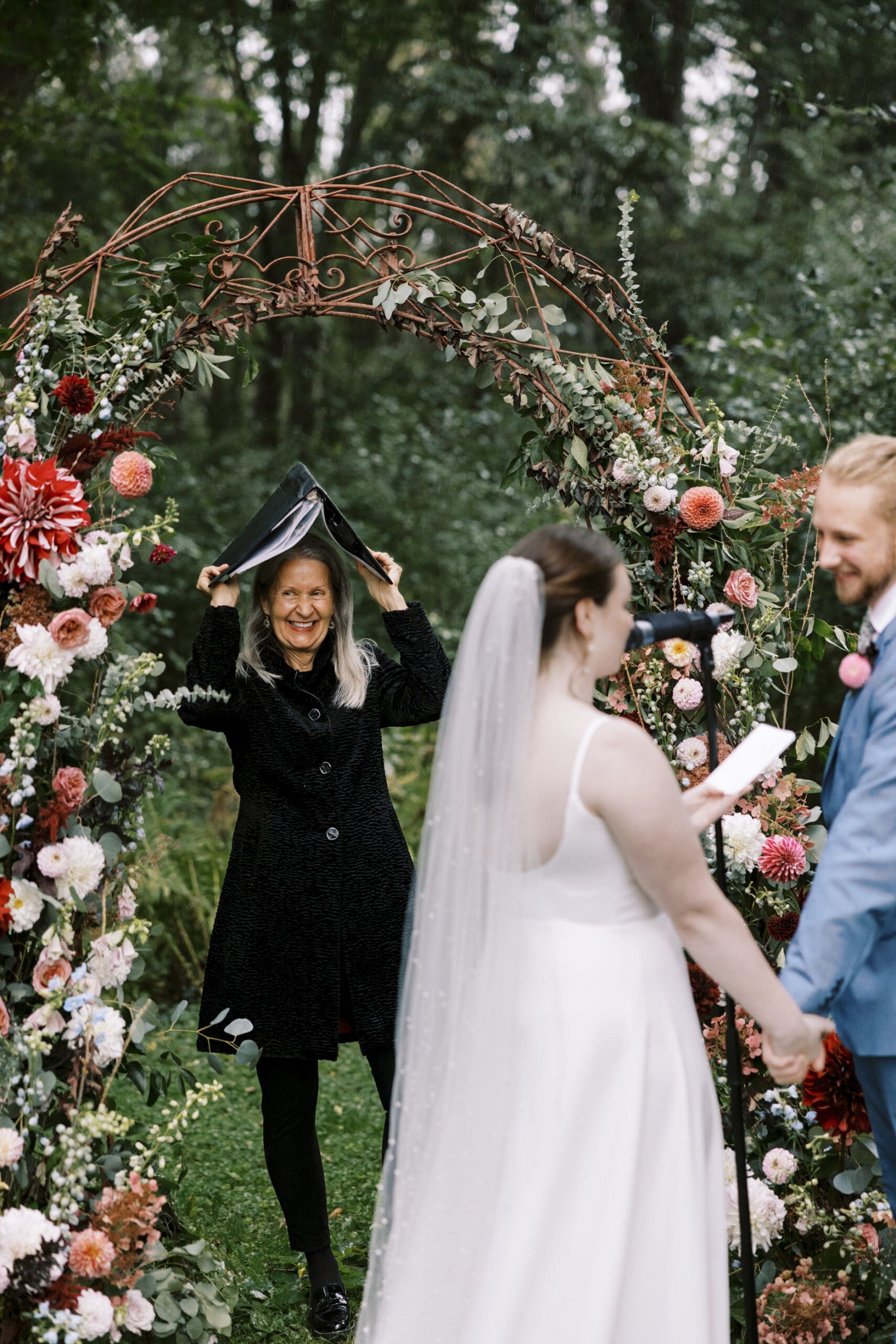 Bride and groom during the rainy garden wedding ceremony at The Outing lodge in Stillwater, Minnesota. Officiant hiding under her binder and smiling while it rains. 